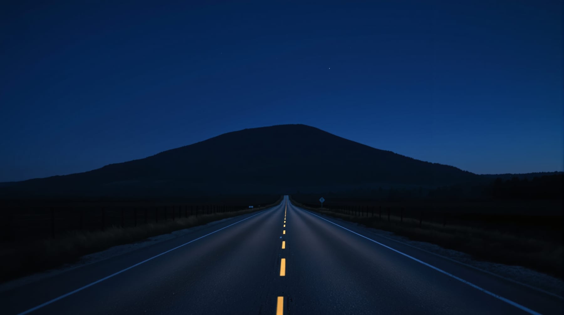 Unlit rural road running through farmland toward a forested mountain at dusk